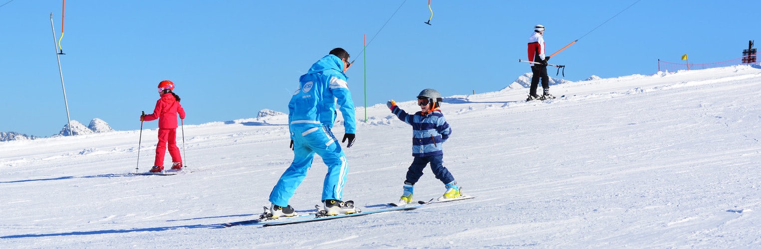 A ski instructor wearing a blue uniform and a young child on a ski lesson in the sun.